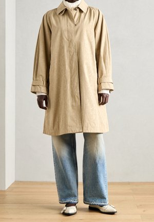 Persoon die een beige trenchcoat, een witte gebreide coltrui, verweerde wijde jeans en witte loafers draagt, staand op een houten vloer.