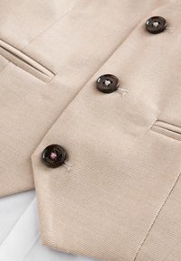 Bež teksturirano tkanje s tamnosmeđim gumbima označenim "Baker" i ušivenim horizontalnim džepovima na odjevnom predmetu.