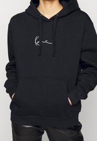 Czarna bluza z kapturem, wykonana z miękkiego materiału, z kieszenią z przodu. Posiada sznurek w kapturze oraz mały biały wzór w okolicy klatki piersiowej.