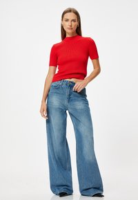 Top corto a maniche corte rosso a coste abbinato a jeans in denim a gamba larga di un blu chiaro. I jeans hanno una finitura leggermente sbiadita e una chiusura con bottone.