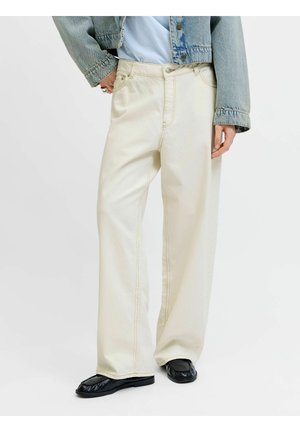 Személy széles szárú krémszínű nadrágot, fekete négyzetorrú cipőket, farmerdzsekit és világoskék felsőt visel, egy kézzel a zsebében áll.