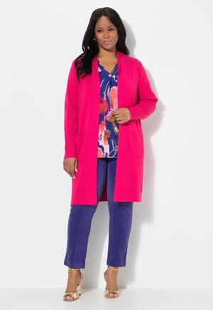 Cardigan long et ouvert de couleur rose vif, porté sur une blouse florale bleu marine, associé à un pantalon bleu retroussé et des sandales à talons dorés.