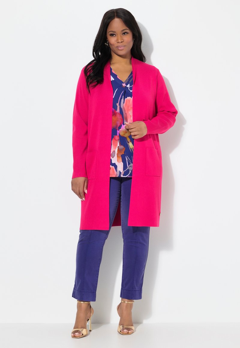 Cardigan lungo aperto di un brillante rosa, indossato sopra una blusa floreale blu navy, abbinato a pantaloni blu con risvolto e sandali con tacco dorati.