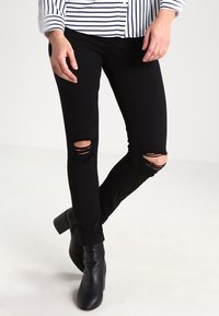 Černé roztrhané džíny s úzkým střihem, s vybledlými oblastmi na kolenou. Spárovány s černými kotníkovými botami na podpatku a bílou pruhovanou košilí s dlouhým rukávem.