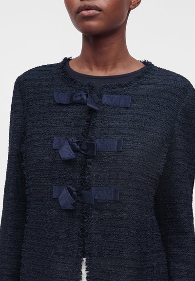 Personne portant une veste bleu marine texturée avec trois nœuds décoratifs bleu marine sur le devant, vue des épaules à la taille sur un fond uni.
