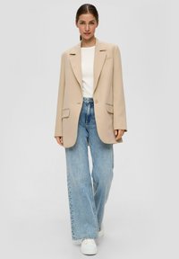 Beige übergroßer Blazer mit Reverskragen, kombiniert mit einem weißen, figurbetonten Oberteil und weiten blauen Jeans. Weiße Sneakers runden das Outfit ab.