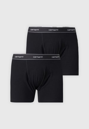 Man zonder hemd met krullend haar, gekleed in zwarte Carhartt boxershorts, staat tegen een effen lichte achtergrond.