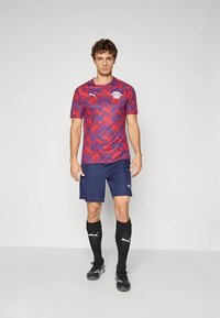 Nogometni dres z rdečim in modrim cikcak vzorcem, kratkimi rokavi, v kombinaciji z mornarsko modrimi kratkimi hlačami ter črnimi nogavicami do kolen. Vidna je znamka Puma.