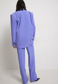 Lavendelfarbenes Anzugset mit einem übergroßen Blazer mit scharfen Schultern und weit geschnittenen Hosen. Glatte Textur und schwarze Knopfakzente an den Ärmeln.