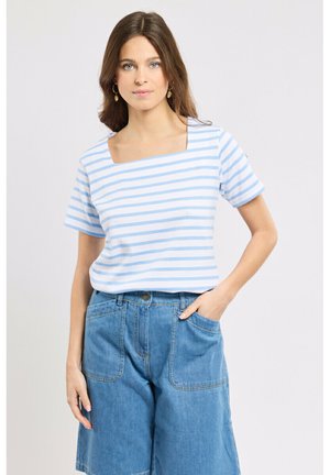 Femme aux cheveux bruns portant un haut à rayures bleues et blanches à encolure carrée et un short en jean bleu ample, main dans la poche, fond neutre.