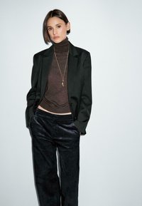 Blazer nero sopra un dolcevita marrone, abbinato a pantaloni in velluto a coste scuri. Le caratteristiche includono una semplice collana d'oro e un design minimalista.