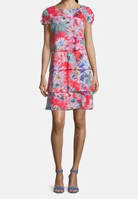 Květinové šaty v červených, růžových a modrých odstínech. Krátké nafouknuté rukávy, vrstvené volány a živé vzory. V páru s modrými sandály na kotníku.