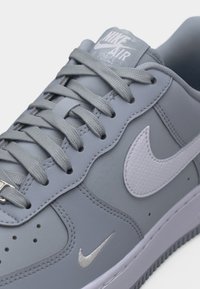Detailný záber na sivú tenisku Nike Air Force 1 so sivými šnúrkami, bielym logom Nike a malými dierkami v oblasti špičky.