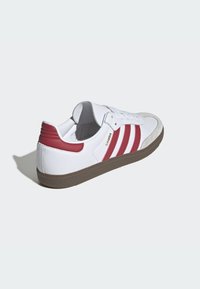 Baskets en cuir blanc avec des accents rouges et trois rayures, dotées d'une semelle en caoutchouc marron et d'un capuchon en suède. Logo "SAMBA" sur le côté.
