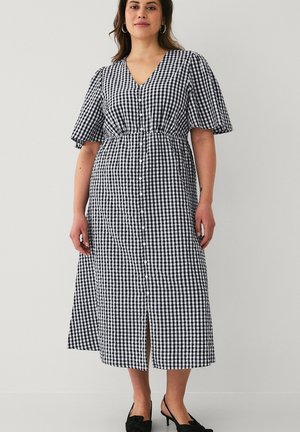 Kvinde iført en sort-hvid ternet midikjole med knapper foran og korte flagrende ærmer, kombineret med sorte slingback hæle.