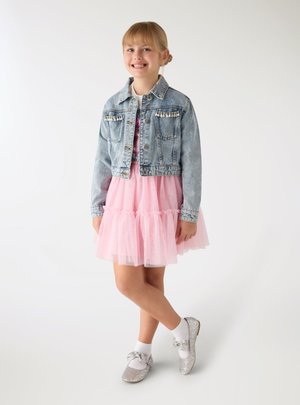 Ragazza sorridente in piedi con le gambe incrociate, indossa un vestito rosa in tulle, una giacca di jeans con decorazioni, calzini bianchi e scarpe glitter argento.