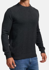 Czarny sweter z teksturowanym wzorem jodełki, okrągłym dekoltem, ściągaczami przy mankietach i dole. Tkanina wydaje się miękka i lekko teksturowana.