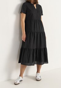 Schwarzes Stufenkleid mit kurzen Ärmeln, V-Ausschnitt und geraffter Struktur an den Stufen, kombiniert mit weißen und schwarzen Sneakern.