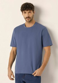 Modrá bavlněná tričko s crew neckem a krátkými rukávy, s uvolněným střihem a hladkou texturou, nošeno přes modré denimové džíny.