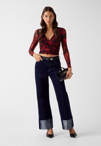 Czerwona, kwiecista bluzka z długim rękawem i dekoltem w kształcie litery V, zestawiona z ciemnoniebieskimi spodnimi spodniami o szerokich nogawkach z denimu. Dodatki obejmują małą czarną torebkę.