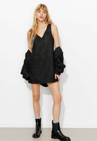 Robe noire sans manches avec un col en V, tissu texturé et coupe ample, associée à des bottines noires et une veste noire surdimensionnée.