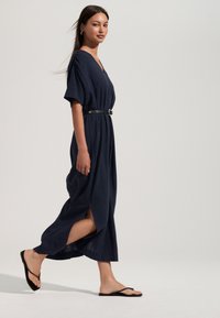 Tumši zila maksi kleita ar īsām piedurknēm, V veida izgriezumu, jostu viduklī un sānu šķēlumiem, valkāta kopā ar melnām flipflop sandalēm. Gluda auduma tekstūra.