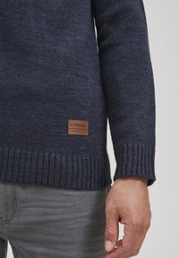 Marineblauer Strickpullover mit strukturiertem Finish, gerippten Bündchen und Saum, mit einem braunen rechteckigen Etikett mit "11 PROJECT" nahe dem Saum.