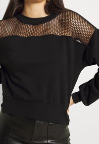Sudadera negra que presenta un panel de malla en los hombros y el escote. El material es suave con puños y dobladillo acanalados, creando una silueta ajustada.