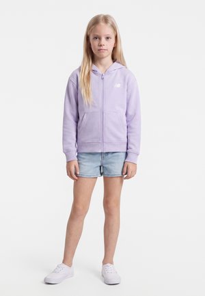 Jeune fille debout, vêtue d'un sweat à capuche zippé violet clair, d'un short en jean bleu clair et de baskets blanches sur un fond blanc.