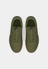 Par nogavica maslinaste boje Nike tenisica s vezicama, prikazana odozgo na bijeloj pozadini.