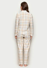 Frau steht barfuß und trägt ein langärmliges Oberteil in Beige, Weiß und Hellblau mit Karomuster sowie passende Hosen, mit dem Rücken zu einer einfarbigen Hintergrund.