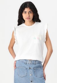 Haut blanc à manches courtes avec détails froncés aux bras et une poche poitrine ornée de pompons colorés. Assorti avec une jupe en jean bleu clair.