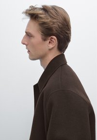 Brun uldfrakke med en afslappet pasform, bred krave og blød tekstur; vist i profil mod en lys baggrund.