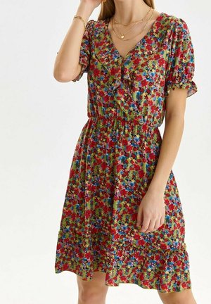 Femme portant une robe fleurie jusqu'aux genoux avec des fleurs rouges, bleues et vertes, un col en V volanté, des manches courtes bouffantes et une taille froncée.