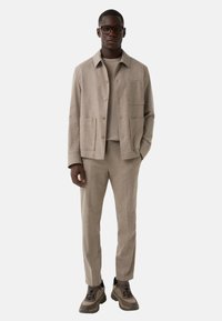 Beige maßgeschneiderte Jacke mit zwei Fronttaschen, getragen über einem passenden leichten Oberteil und eng anliegenden Hosen. Akzentuiert mit braunen Turnschuhen.