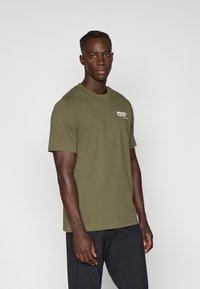 Camiseta de algodón verde oliva con mangas cortas, cuello redondo y un pequeño logotipo blanco en el lado izquierdo del pecho. Textura suave y diseño simple.