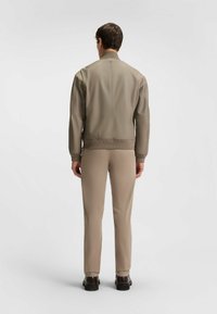 Ljus taupe zip-up jacka med en figursydd design och ribbad nederdel, kombinerad med beiga byxor och svarta skor, sedd bakifrån.