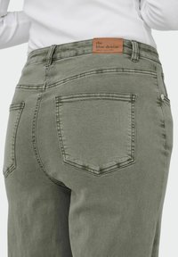 Gros plan d'une personne portant un jean en denim vert olive avec des poches arrière et une étiquette de marque en cuir beige portant l'inscription "the blue denim".