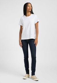 Camiseta blanca de manga corta combinada con vaqueros skinny oscuros y zapatillas beige; el modelo está de pie con los brazos relajados a los lados y una expresión neutral.