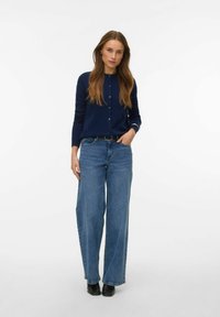 Tmavě modrý propínací svetr s dlouhými rukávy, kombinovaný s širokými světle modrými džínami a černými kotníkovými botami. Jednoduchý a klasický design.