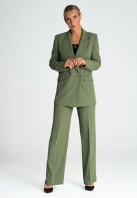 Oliwkowy garnitur szyty na miarę z podwójnym zapięciem, posiadający klapy i szerokie spodnie, zestawiony z czarnymi szpilkami.
