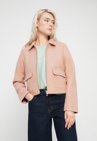 Ανοιχτό ροζ κοντομάνικο σακάκι από μαλακό ύφασμα, με φερμουάρ εμπρός, επίπεδη γιακά και δύο μπροστινές τσέπες. Συνδυασμένο με πράσινο τοπ και τζιν παντελόνι.