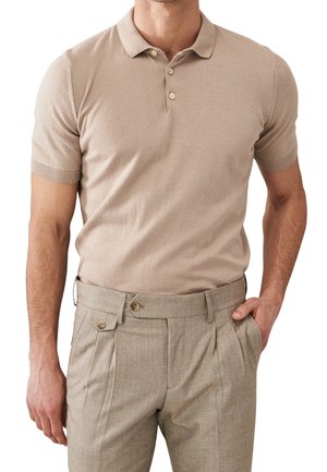Uomo che indossa una polo beige a maniche corte infilata in pantaloni beige a vita alta con pieghe e tasche con bottoni, con una mano in tasca.