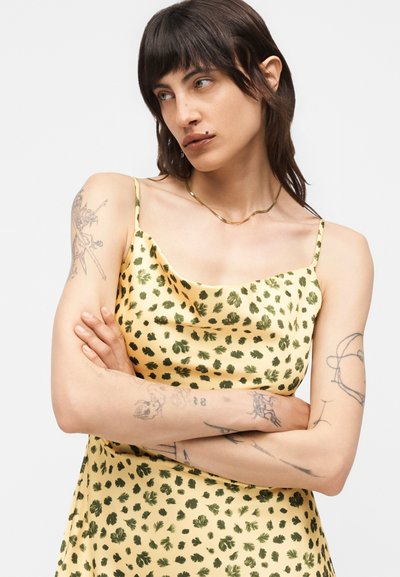 Άτομο με σκούρα μαλλιά και τατουάζ που φοράει κίτρινο φόρεμα με πράσινα σχέδια φύλλων, χέρια σταυρωμένα, κοιτάζει προς τα πλάγια σε λευκό φόντο.