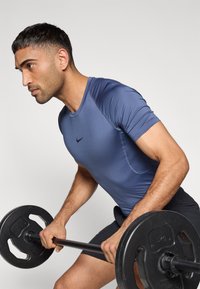 Κοντομάνικο αθλητικό πουκάμισο σε ναυτικό μπλε με εφαρμοστή γραμμή, που διαθέτει αντίθετο μαύρο λογότυπο στο στήθος, συνδυασμένο με σκούρο σορτς και μια μπάρα με βάρη.