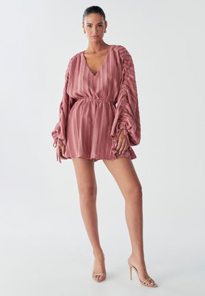 A model poses in a pink, textured dress with voluminous sleeves and an elastic waist, paired with nude high-heeled sandals against a light background.