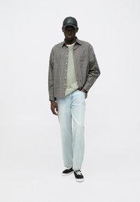 Camicia a quadri marrone e beige, maglione girocollo verde, jeans azzurri e scarpe da ginnastica nere; indossa anche un berretto nero.