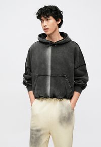 Jovem com cabelo encaracolado vestindo um capuz preto e cinza e calças creme com manchas de tinta preta, mãos nos bolsos, olhando de lado.