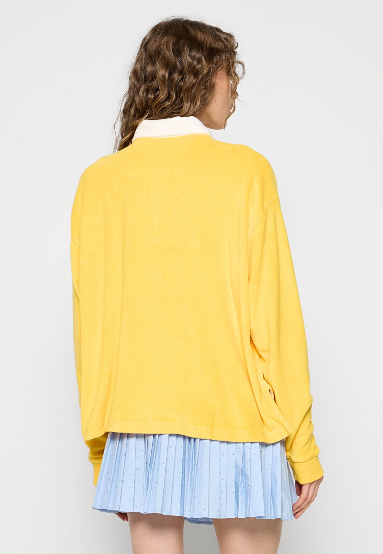 Felpa gialla oversize con maniche lunghe, caratterizzata da un colletto bianco e una texture morbida. Abbinata a una gonna plissettata azzurro chiaro.