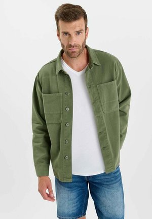 Homme portant une chemise verte boutonnée par-dessus un T-shirt blanc et un short en jean, debout devant un fond clair uni.